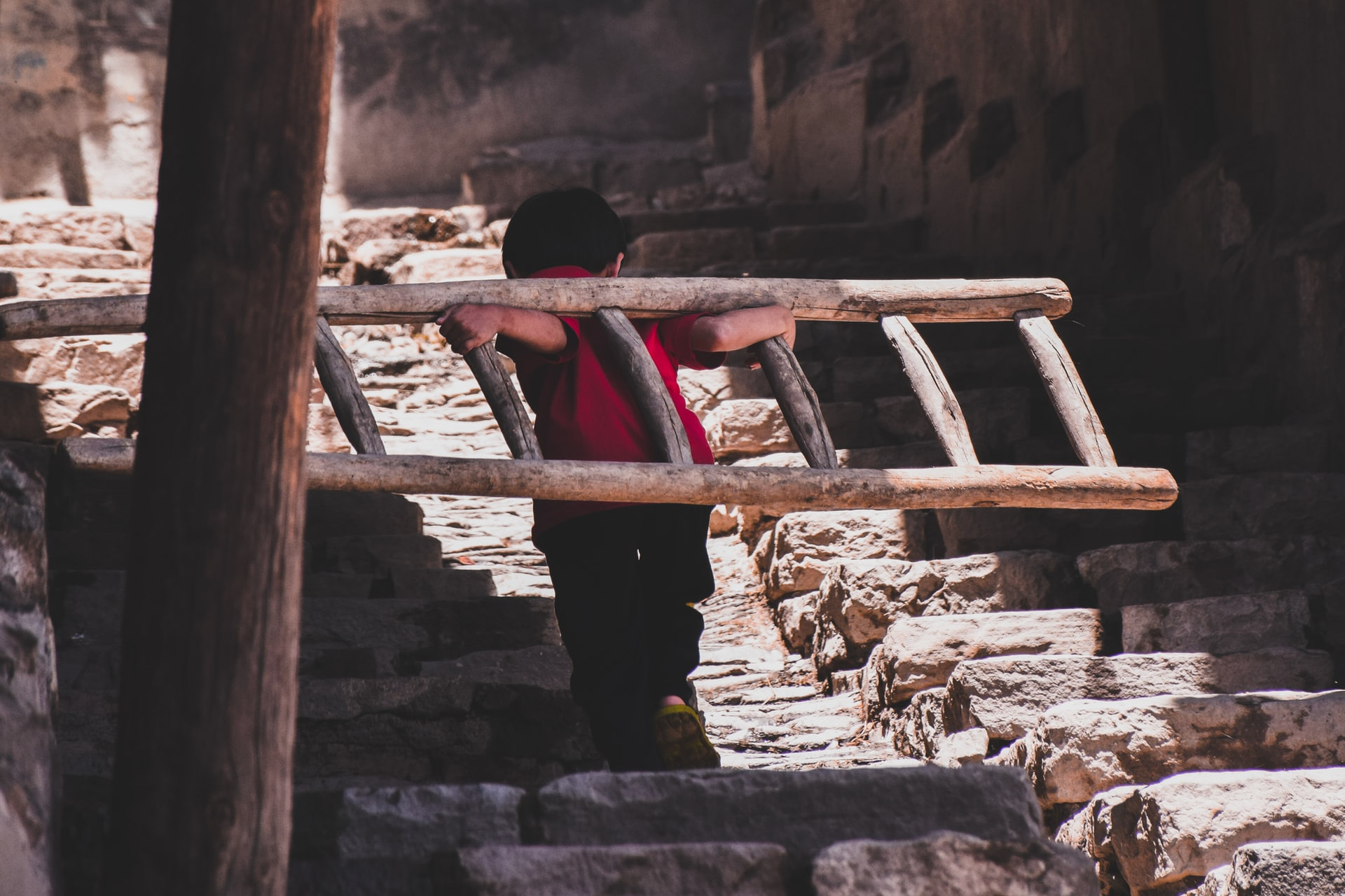 Child holds a ladder across shoulders while walking up rough steps
