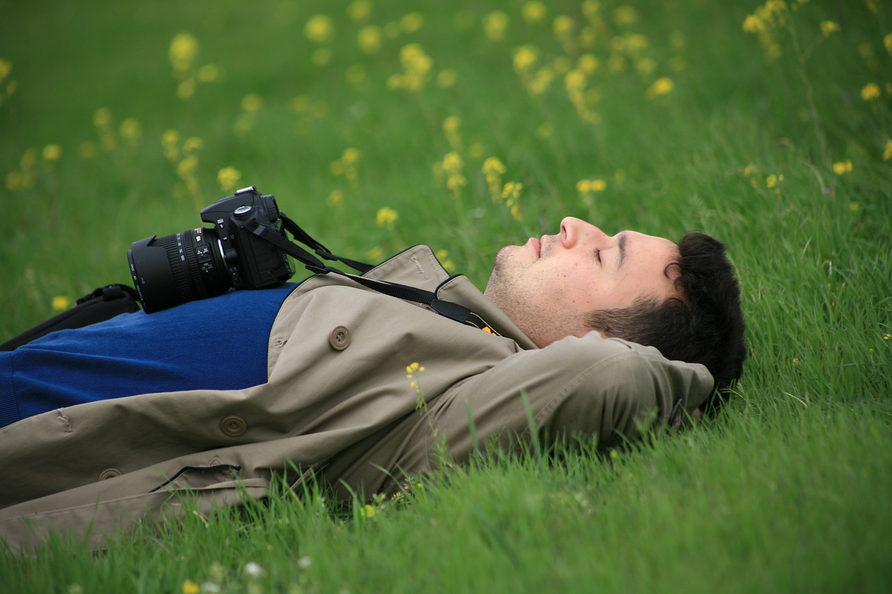 Photographer lying on grass among yellow flowers, DSLR camera hanging from his neck, arms behind his head