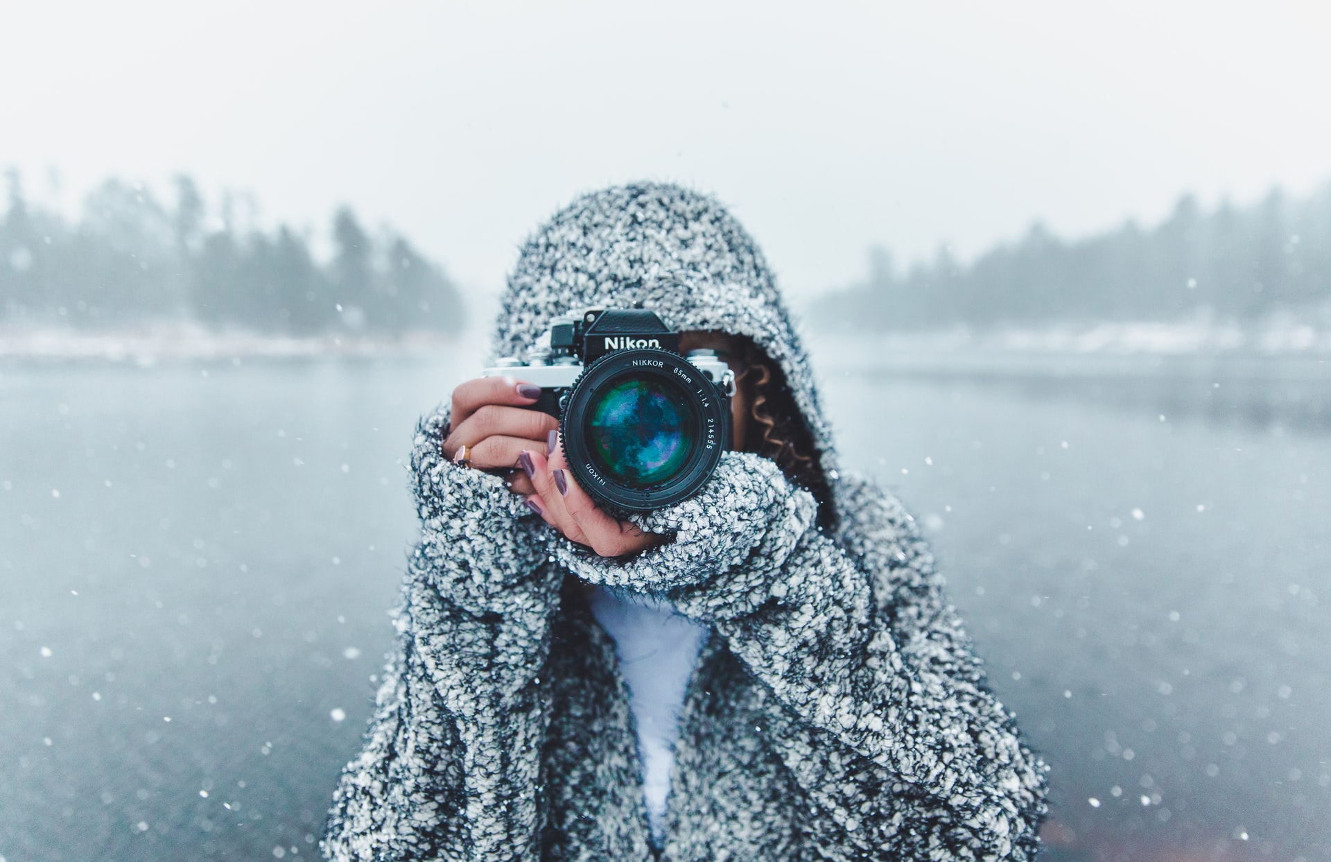 Photographer wrapped up warm and pointing camera at the viewer, standing in front of a lake surrounded by trees, snow falling all around
