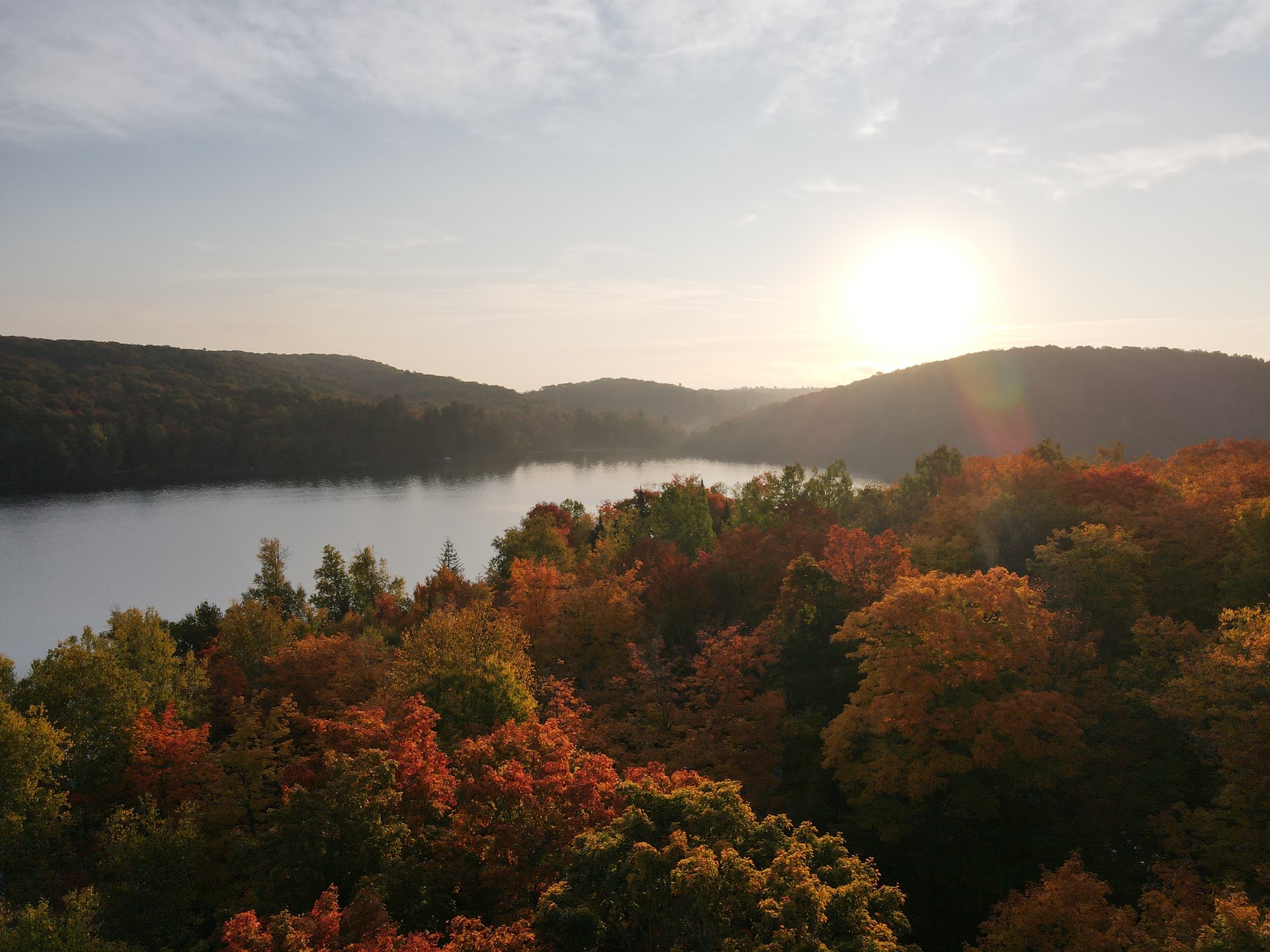 Scenic photo of trees beside a lake displaying autumn colours, hills in background with sun cresting the peak