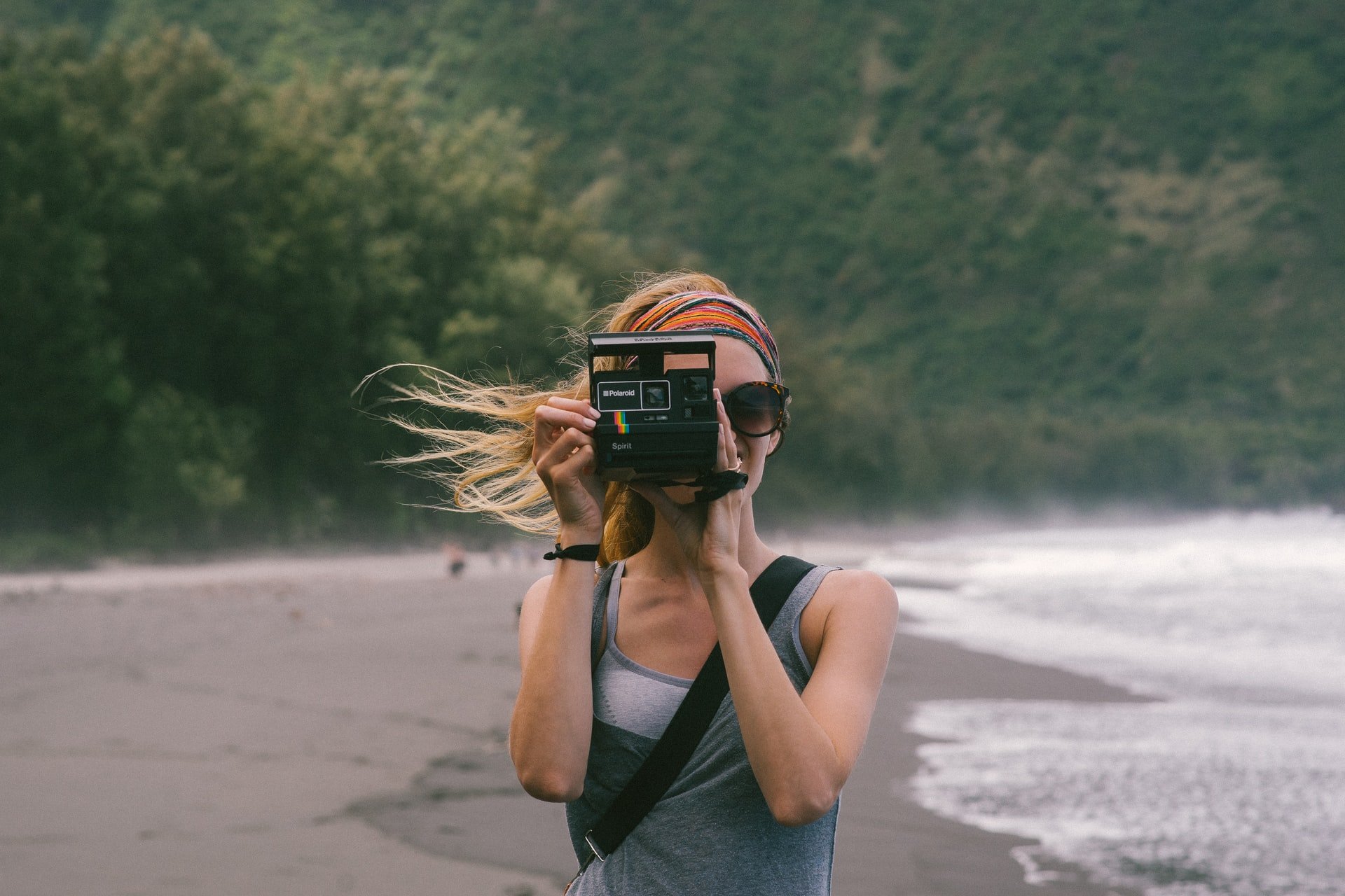 Photographer standing on a beach, trees in background, tide coming in, holding a Polaroid camera pointed at the viewer