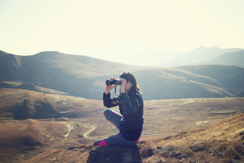 woman crouched down amidst rolling hills, looking through binoculars